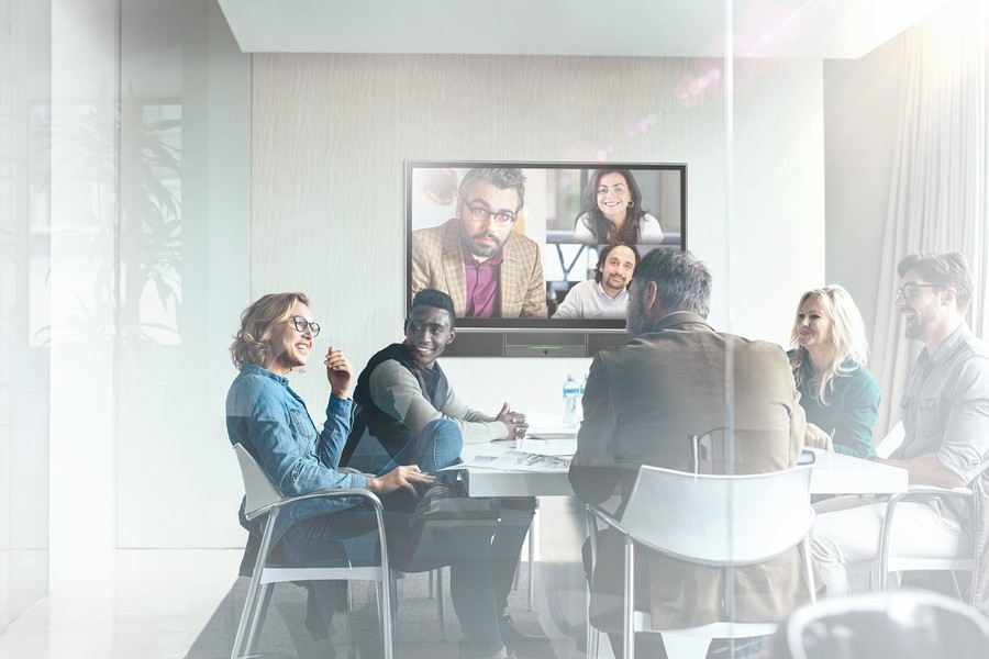 Room of employees in a conference room with video conferencing on the display screen at front of room