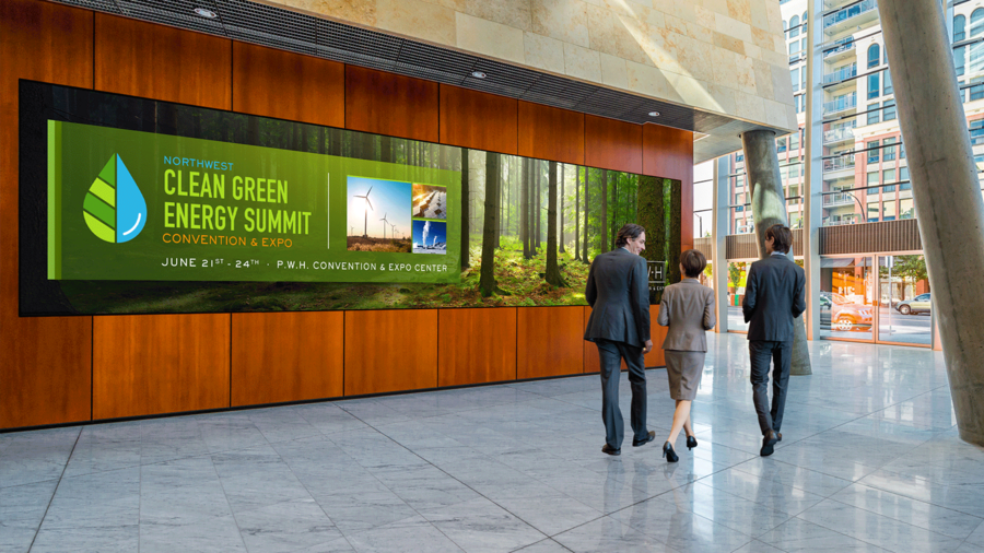 A wall-sized digital signage display advertises a green energy summit. Three office workers walk by the sign. 