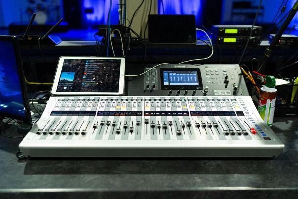 A mixing console with various sliders, buttons, and screens. An iPad and other devices are connected and placed on the console.