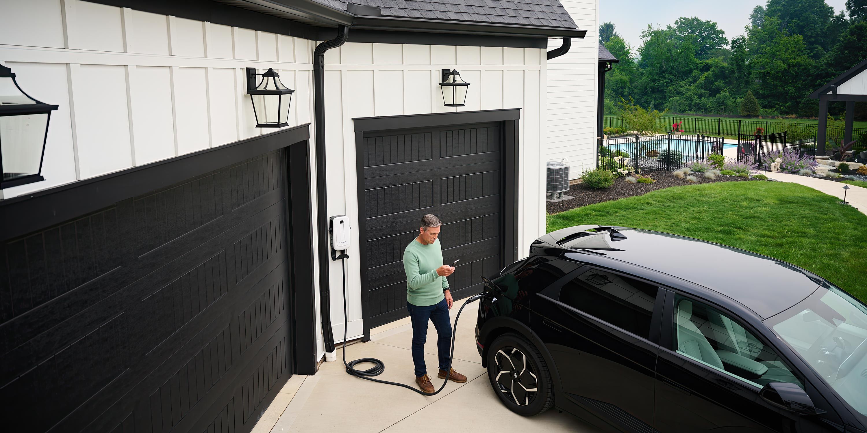 A man charging his electric vehicle outside a modern home with black garage doors, using a wall-mounted charger while checking his phone.