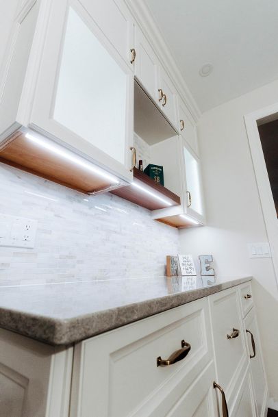 A close-up of a kitchen countertop with white cabinets above. The cabinets have brass handles and under-cabinet LED lighting, with a few decorative items placed on the counter.