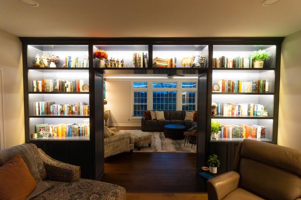 A cozy reading nook with built-in bookshelves framing an entryway. The shelves are filled with books and decorative items, and the room beyond features a comfortable seating area with large windows.