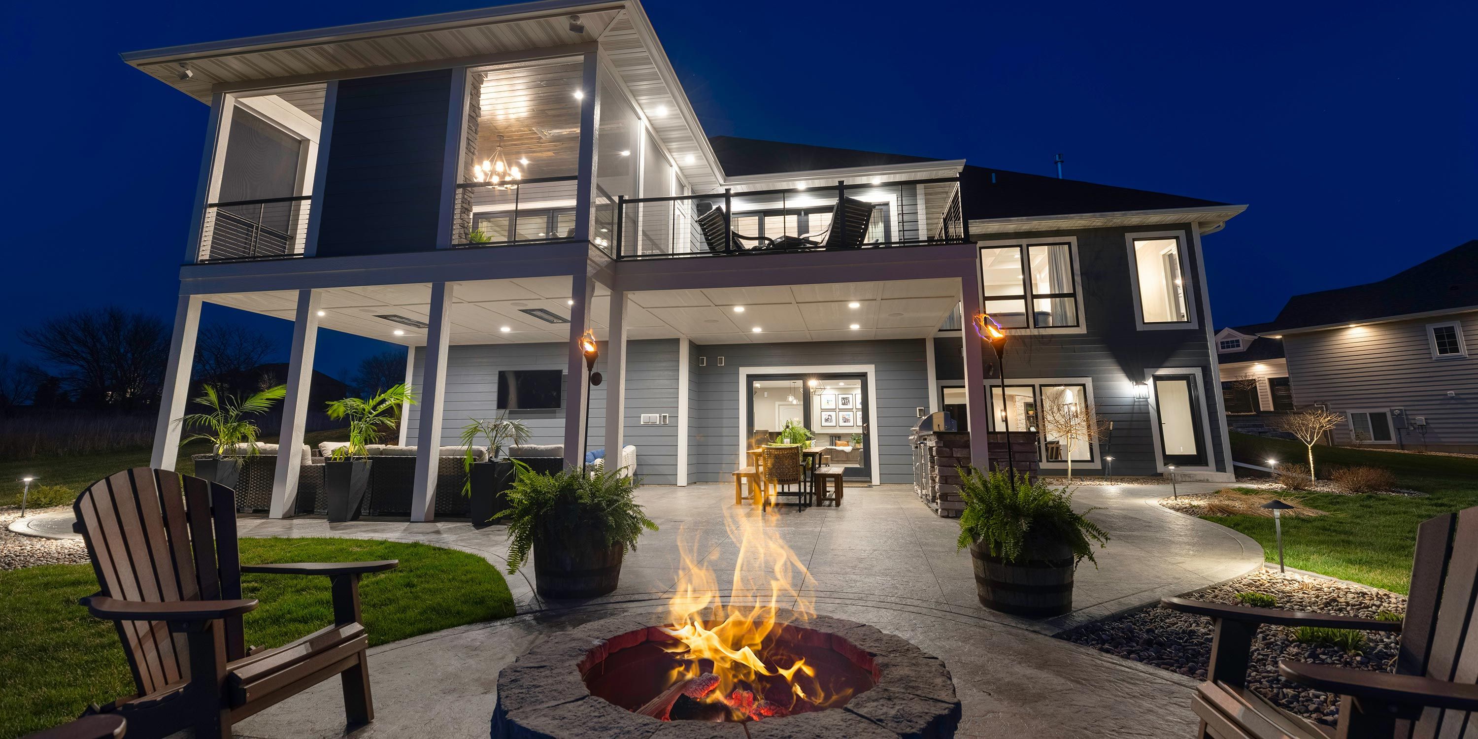 A beautifully lit two-story home with an outdoor living space, fire pit, and cozy seating area, creating a warm and inviting backyard atmosphere at night.