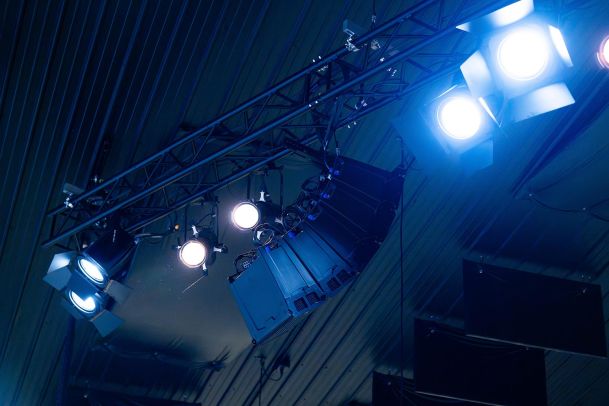 A close-up view of stage lights and speakers mounted on a truss, with blue and white lights shining down.