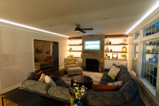 A living room with a stone fireplace, a mounted TV, and built-in shelves displaying decorative items. The room has a U-shaped sectional sofa, an armchair, and a coffee table, creating a warm and inviting space.