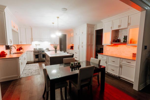 A spacious kitchen and dining area with white cabinetry and modern LED lighting. The cabinets have a warm underglow, and the dining table is set for a meal, creating a welcoming atmosphere.