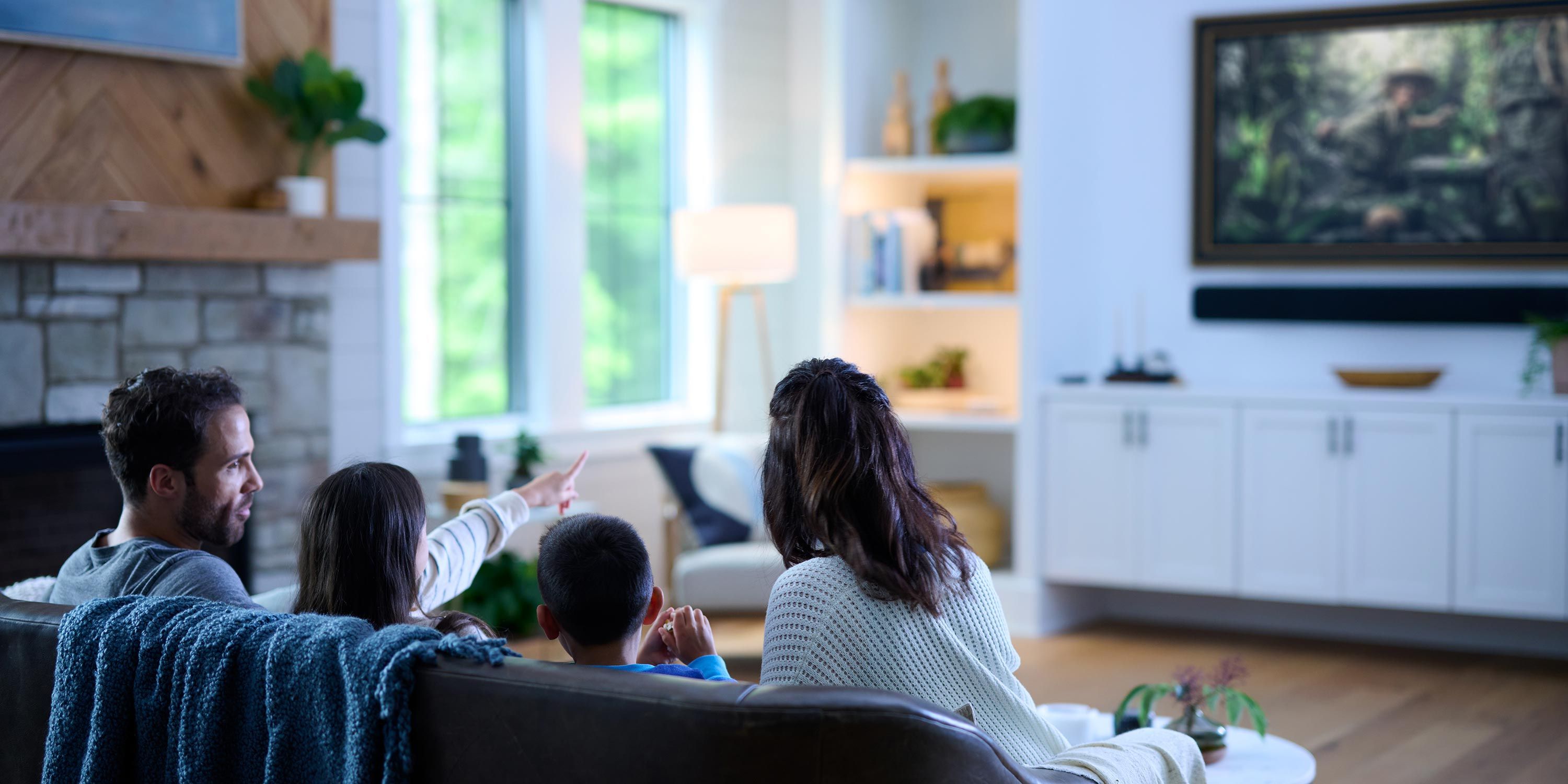 A family sitting on a cozy couch in a living room, pointing to a wall-mounted television displaying a scenic image.