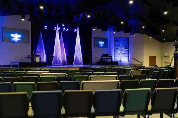 A church auditorium with rows of empty chairs facing a stage. The stage has a drum set, music stands, and tall, thin, conical decorations. Two large screens display a "Welcome" message.
