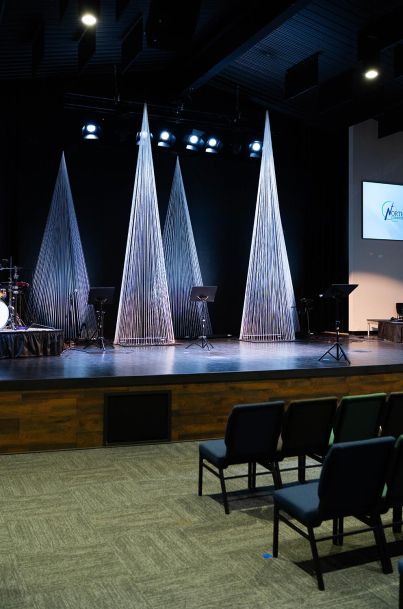 The stage in the auditorium with tall, thin, conical decorations and music stands. The left side of the stage has a drum set.