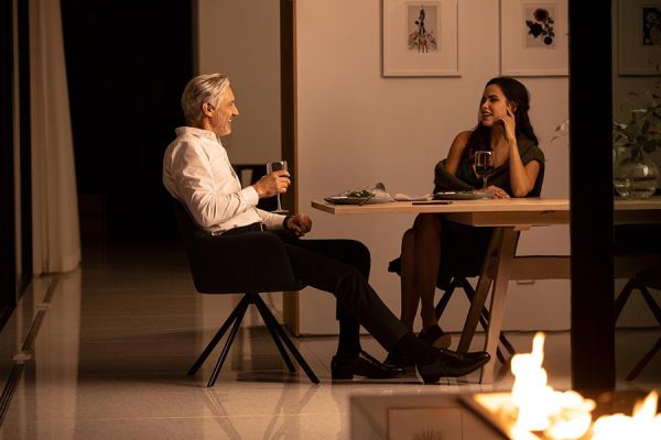 A cozy dining scene with two people enjoying wine at a table, with ambient firelight in the foreground.