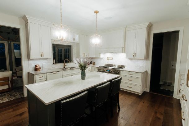 A modern kitchen with white cabinets, a marble countertop island, and stainless steel appliances. The kitchen is well-lit with two pendant lights hanging above the island and has a bouquet of flowers as a centerpiece.