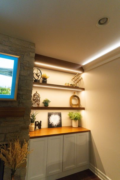 A close-up view of built-in shelves in a living room, decorated with various ornaments, plants, and a framed piece of artwork. The shelves are lit with soft lighting, highlighting the decor.