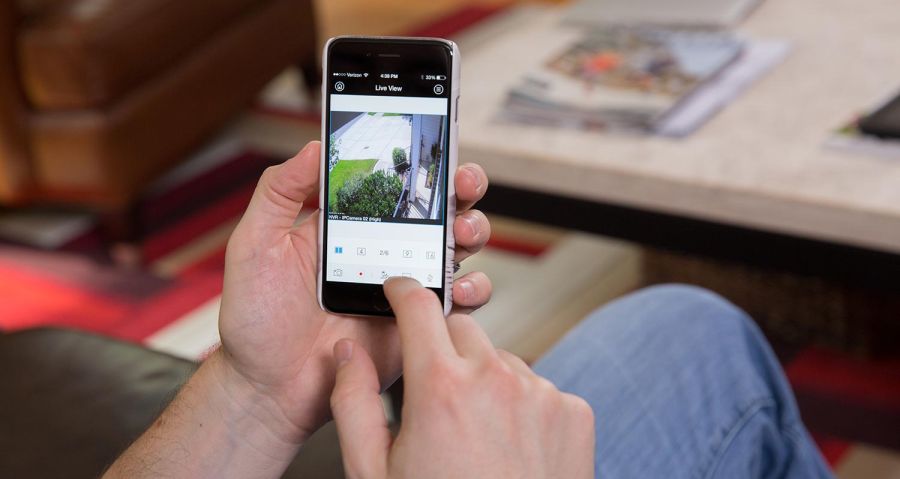 A person holding a smartphone, monitoring a live security camera feed that shows an outdoor area with a pathway and bushes.